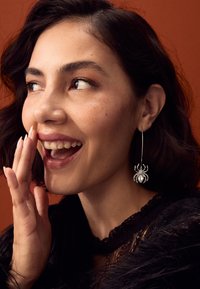 Silver spider-shaped earrings with rhinestone accents, worn by a person with dark hair, against a warm orange background.
