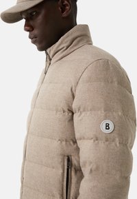 Man in een beige gewatteerde jas met een ritszak en een bijpassende beige pet, naar links gekeerd tegen een neutrale achtergrond.