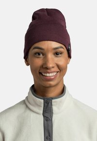 Maroon knit beanie with a ribbed texture, featuring a small black tag. Worn with a white fleece jacket with gray accents and buttons.