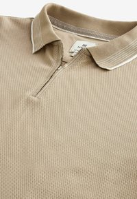 Polo beige texturé à manches courtes avec col présentant des rayures blanches et une demi-fermeture éclair à l'avant.