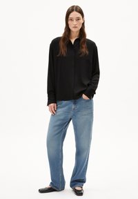 Schwarzes langärmliges Hemd mit Knöpfen und Kragen, getragen mit hellblauen weiten Jeans und schwarzen flachen Schuhen. Glatter Stoff und lockere Passform.