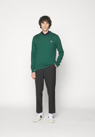 Green knitted sweater with long sleeves and a round neckline, paired with black trousers and white sneakers. Small logo on the sweater.
