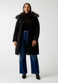 Cappotto imbottito nero con zip frontale, dotato di cappuccio foderato in pelliccia. Abbinato a jeans a vita alta blu a zampa d'elefante e stivaletti neri a punta.