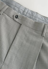 Gros plan d'un pantalon en tissu à carreaux avec un bouton noir, une boucle de ceinture et une ceinture intérieure présentant un motif chevron.