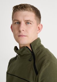 Olivově zelený fleecový svetr s vysokým límcem, který má polo-zip zapínání a černé akcentové švy podél ramen.