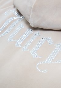Gros plan sur un tissu beige orné de strass formant un texte cursif stylisé partiellement visible.