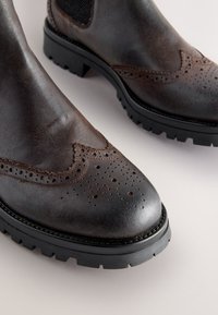 Dunkelbraune Leder-Stiefeletten mit Brogue-Details, leicht spitz zulaufender Spitze und robuster schwarzer Gummisohle. Strukturierte Oberfläche in der Nähe der Schnürsenkel.