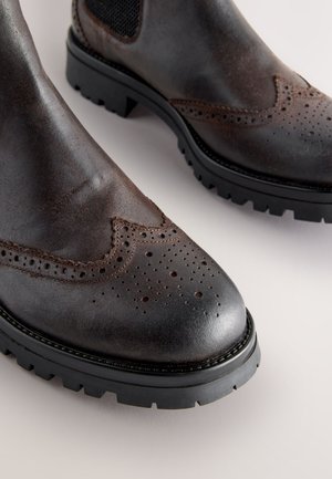 Dunkelbraune Leder-Stiefeletten mit Brogue-Details, leicht spitz zulaufender Spitze und robuster schwarzer Gummisohle. Strukturierte Oberfläche in der Nähe der Schnürsenkel.