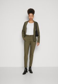 Olivově zelený přizpůsobený blazer a ladící slim-fit kalhoty, doplněné bílým topem a černými kotníkovými botami. Jednoduchá kabelka jako akcent.