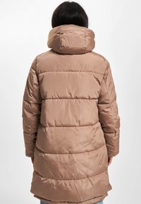 Person med en lang, polstret, beige vinterfrakk med høy krage, sett bakfra mot en ensfarget hvit bakgrunn.