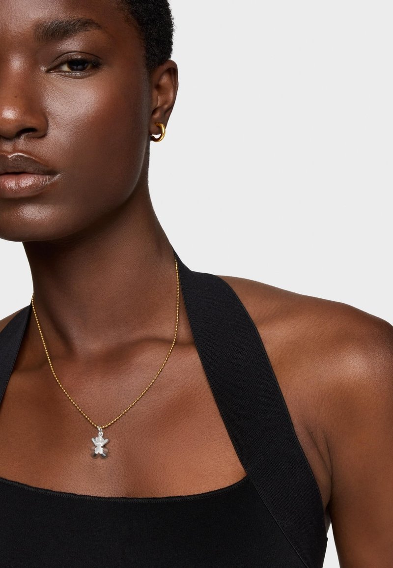 Collana in oro con un piccolo ciondolo a forma di orso in argento. Il modello indossa una maglietta nera senza maniche con spalline larghe. Texture della pelle liscia.