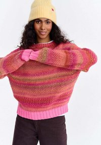 Kobieta z długimi kręconymi włosami, ubrana w żółtą czapkę typu beanie, wielokolorowy różowo-pomarańczowy sweter w paski i ciemne spodnie, na jednolitym białym tle.