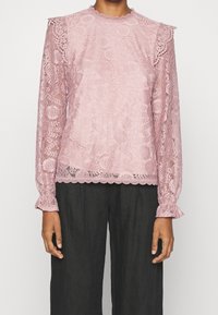 Top lungo a maniche con pizzo rosa, con scollatura e bordi delle maniche a scallop, caratterizzato da intricate fantasie floreali e geometriche. Indossato con pantaloni scuri.