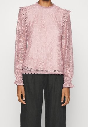 Top lungo a maniche con pizzo rosa, con scollatura e bordi delle maniche a scallop, caratterizzato da intricate fantasie floreali e geometriche. Indossato con pantaloni scuri.