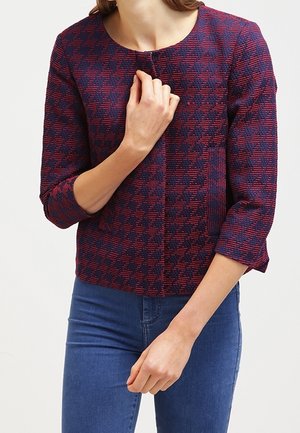 Vrouw die een rood en marineblauw pied-de-poulemotief jasje draagt met driekwart mouwen en een blauwe spijkerbroek, en die het jasje bij de kraag vasthoudt.