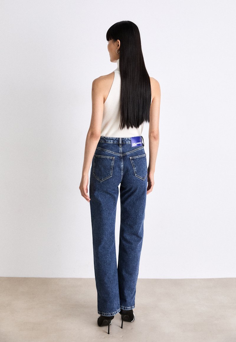 Hoch taillierte blaue Denim-Jeans mit einer lockeren Passform, die über Gesäßtaschen und ein lila Etikett am Bund verfügt, getragen mit einem ärmellosen Oberteil.
