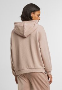 Lys rosa hettegenser med en romslig silhuett, snorhette og ribbestrikket kant. Materialet ser mykt ut med en subtil tekstur.