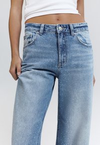 Kobieta w jasnoniebieskich, wysokich spodniach dżinsowych z małymi kryształkami i białym krótki topem, trzymająca boczny szew spodni.