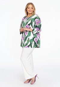 Grüne Blumen-Tunika mit lila und weißen Mustern; kombiniert mit weißen Hosen und lila Heels. Sie hat einen lockeren Schnitt und Dreiviertelärmel.
