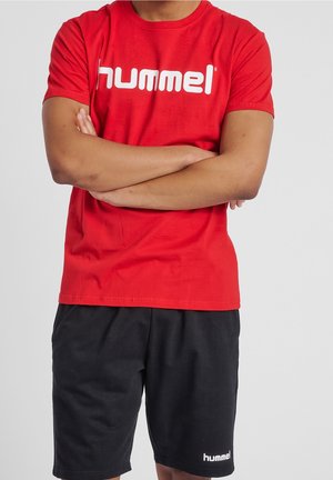 Personne portant un t-shirt rouge Hummel, les bras croisés, et un short noir Hummel, sur un fond uni.