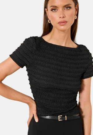 Woman with long brown hair wearing gold hoop earrings, a textured black short-sleeve top, and black pants with a slim belt.