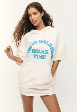 Jeune femme aux longs cheveux portant une robe sweat à capuche blanche avec un texte bleu « Life in Balance Relax Time », tenant un cordon de capuche et posant.