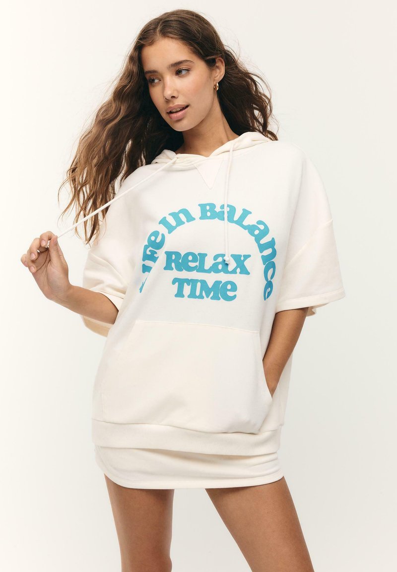 Jeune femme aux longs cheveux portant une robe sweat à capuche blanche avec un texte bleu « Life in Balance Relax Time », tenant un cordon de capuche et posant.