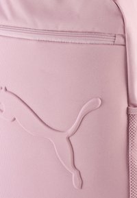 Mochila de tela rosa claro con un logo de Puma texturizado, un bolsillo con cremallera y detalles de malla en los laterales.