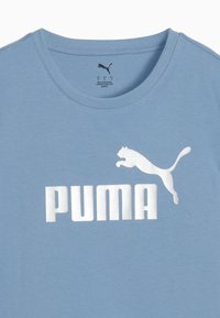 Šviesiai mėlynas medvilninis marškinėlis su baltu PUMA logotipu ir šuoliuočiu katinu. Apvalus kaklas, trumpomis rankovėmis, lygiai švelni tekstūra.