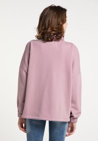 Túlnagy rózsaszín pulóver, magas gallérral és bordázott mandzsettákkal, sima textúrával és cropped hosszal, farmerral viselve.