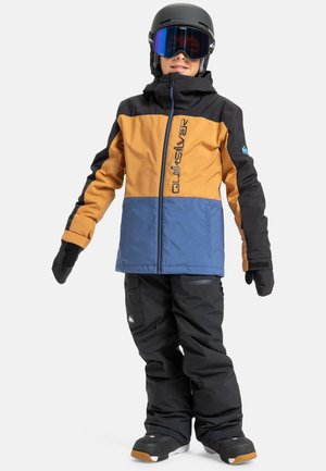 Enfant portant une veste de neige colorée en blocs de marron, bleu et noir, un pantalon de neige noir, des gants, un casque et des lunettes de ski, se tenant sur fond blanc.