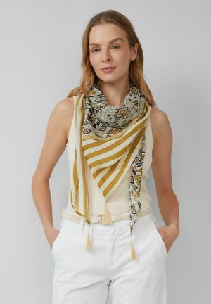 Frau trägt ärmelloses elfenbeinfarbenes Top und weiße Hose, accessoriert mit einem beigen und gold gemusterten Schal mit Quasten um den Hals.