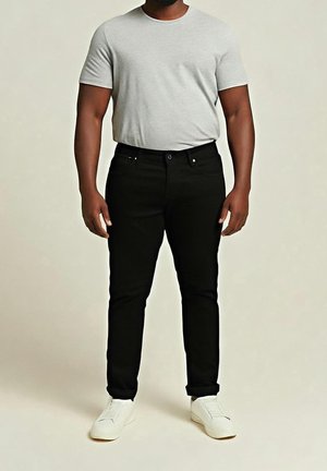 Mann trägt ein hellgraues T-Shirt mit kurzen Ärmeln, schwarze Jeans und weiße Turnschuhe und steht vor einem schlichten beigefarbenen Hintergrund.