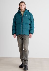 Man wearing teal puffer jacket, beige shirt, gray jeans, and black shoes, standing against a plain white background.