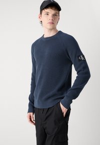 Maglione lavorato a maglia blu navy con polsini e orlo a coste, con una toppa con logo sulla manica. Indossato con pantaloni cargo neri, tasca laterale visibile.