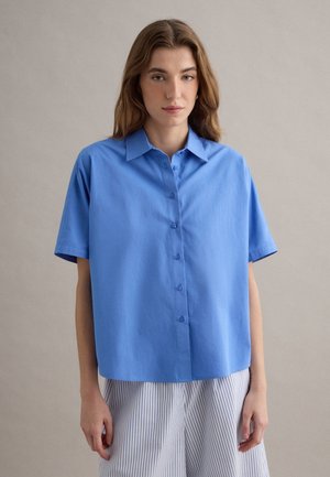 Mujer vistiendo una camisa azul de manga corta y suelta con botones y pantalones blancos con finas rayas verticales, posando frente a un fondo liso.