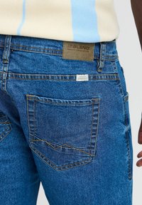 Jeans de denim azul con un acabado texturizado, que cuentan con bolsillos traseros cosidos, hilo dorado de contraste y una etiqueta de cuero sobre la cinturilla.