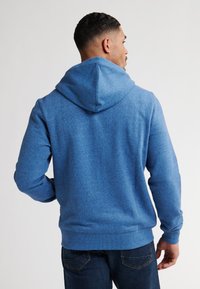 Felpa con cappuccio blu dal tessuto morbido, con polsini aderenti e tasca a marsupio. Indossata con jeans scuri, che mostrano un design casual.