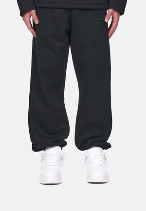 Schwarze Sweatpants, lockere Passform, mit strukturierter Oberfläche. Kombiniert mit weißen Sneakers, die eine glatte, abgerundete Zehe und Schnürung aufweisen.