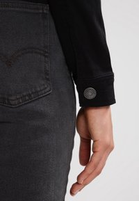 Czarna koszula dżinsowa z ozdobnym mankietem, z metalowym guzikiem. Dżinsy w ciemnoszarym kolorze z widocznym wzorem tylnej kieszeni.