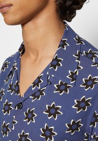 Gros plan d'une personne portant une chemise bleue avec un motif floral noir et blanc et un col, mettant l'accent sur le cou et le haut de la poitrine.