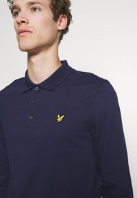 Mornarski polo majica dugih rukava s teksturiranim materijalom, dvobojni gumbasti ovratnik i izvezena žuta ptica na lijevoj strani prsnog koša.