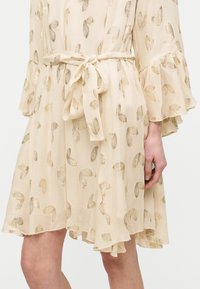 Μπεζ φόρεμα με μακριά μανίκια, μανσέτες με βολάν, κέντημα με φύλλα χρυσού και δεμένη υφασμάτινη ζώνη στη μέση.