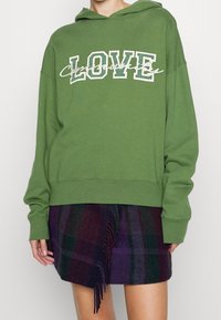 Groene hoodie met "LOVE" in het wit en zwart erop gedrukt, met een relaxte pasvorm en lange mouwen. Draag deze met een paars geruit rokje met franjes.