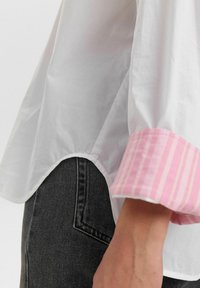 Camicia bianca con polsini a righe rosa, caratterizzata da un orlo curvo. Il tessuto sembra liscio e leggero, abbinato a jeans grigio scuro.