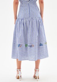 Nainen, jolla on yllään sinivalkoraidallinen midi-hame, jossa on brodeerattuja kukkakuviota helmassa, yhdistettynä valkoisiin korkokenkiin.