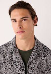 Camicia a maniche corte con motivo paisley bianco e nero e colletto appuntito. Indossata sopra una t-shirt verde scuro tinta unita. Tessuto dalla texture liscia.