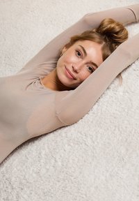 Lange mouwen, beige bodysuit met een aansluitend ontwerp en zachte, rekbare stof die de contouren accentueert, gedragen door een persoon tegen een gestructureerde achtergrond.