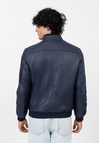 Veste en cuir bleu marine avec un col montant, des poignets côtelés et une texture lisse. Elle présente des coutures détaillées et une poche sur le côté de la manche.