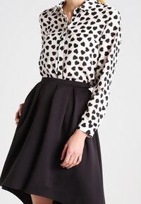 Langärmlige Bluse in Elfenbein mit schwarzem Herzmuster, kombiniert mit einem schwarzen Faltenrock mit asymmetrischem Saum. Glatte Stofftexturen.